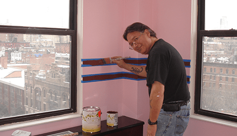A man wearing glasses, smiling while painting a wall pink
